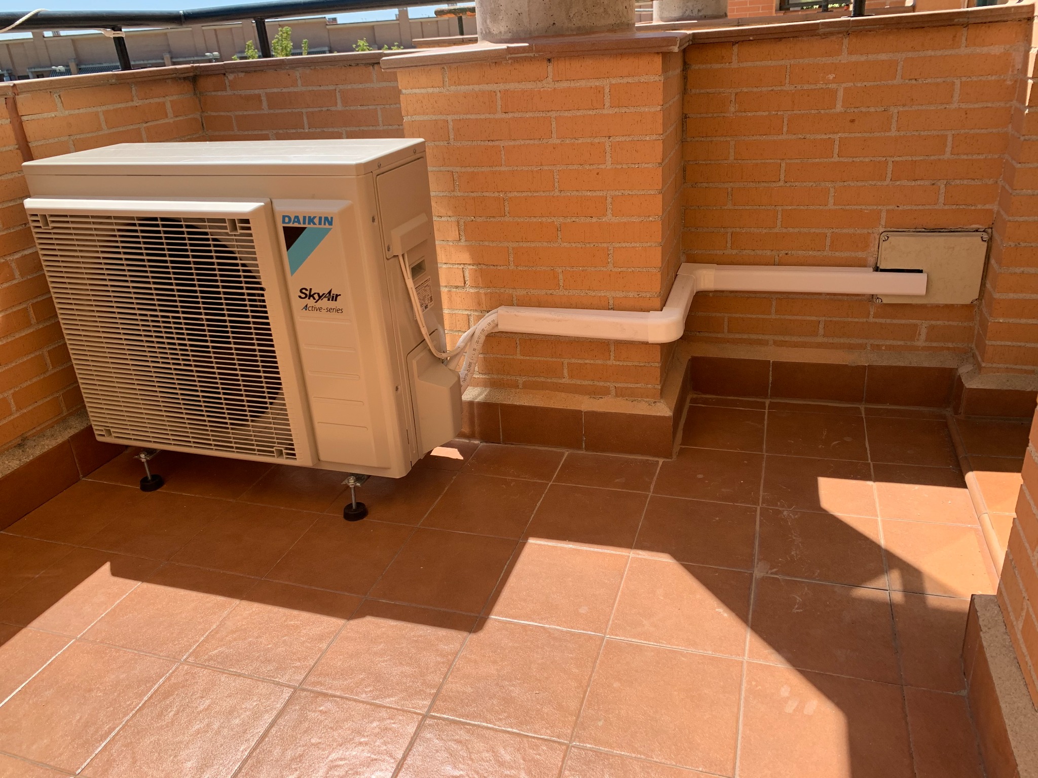 Unidad exterior de aire acondicionado instalada en una terraza con canalización sobre pared de ladrillo.
